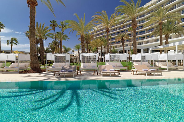 Hotelgebäude mit mehreren Stockwerken hinter einer Poollandschaft mit Liegen und Palmen.