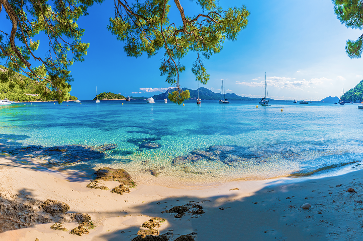 Ein sonniger Sandstrand mit klarem Wasser und mehreren Booten, die sanft an der Küste liegen.