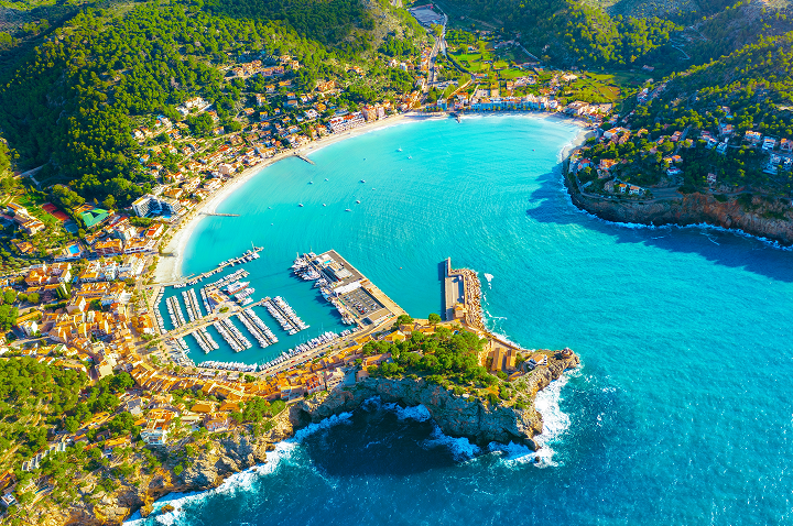 Luftaufnahme eines schönen Strandes und Hafens in Mallorca, umgeben von klarem Wasser und grünen Hügeln.