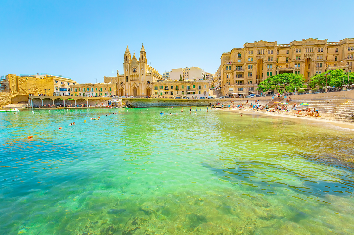Strand in Valletta, Malta, mit klarem Wasser und historischen Gebäuden im Hintergrund.