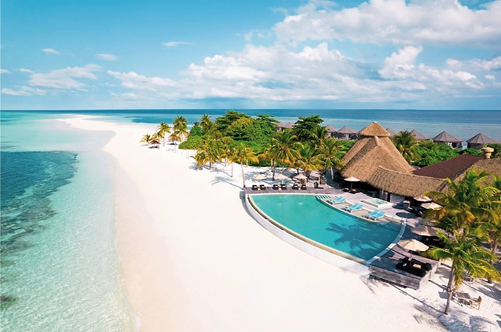 Strandresort mit Pool, Palmen und Strohdachbungalows an weißem Sandstrand und türkisfarbenem Meer unter blauem Himmel mit Wolken.
