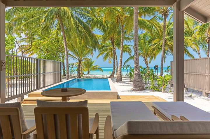 Blick von einer überdachten Terrasse mit Liegen und Stühlen auf einen Pool, Palmen und das Meer im Hintergrund.