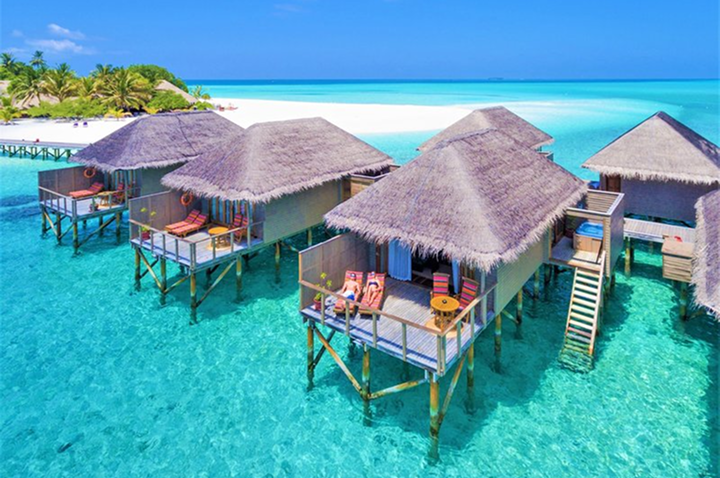 Mehrere strohgedeckte Bungalows auf Stelzen stehen im türkisfarbenen Wasser vor einem weißen Sandstrand mit Palmen.