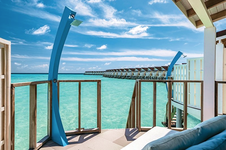 Blick von einer Terrasse mit blauem Außenduschelement auf türkisfarbenes Meer und eine Reihe von über dem Wasser gebauten Bungalows unter blauem Himmel mit Wolken.
