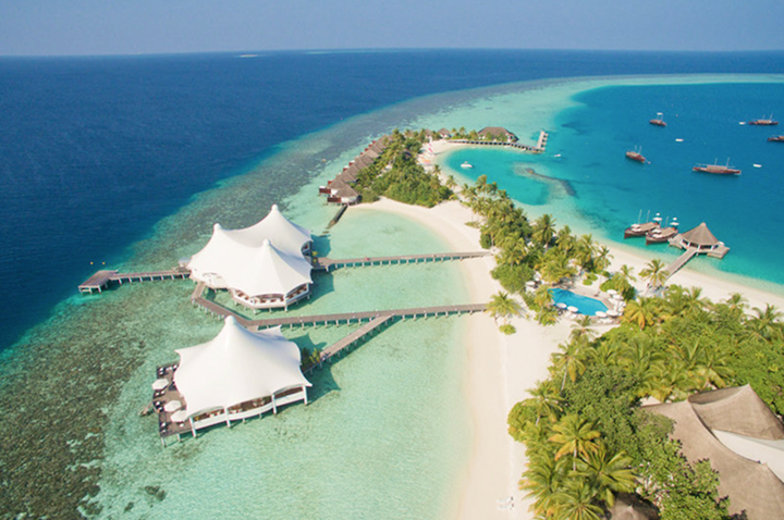 Luftaufnahme einer tropischen Insel mit Sandstrand, Palmen, zwei großen weißen Zeltdächern über Wasser und mehreren Booten im türkisfarbenen Meer.
