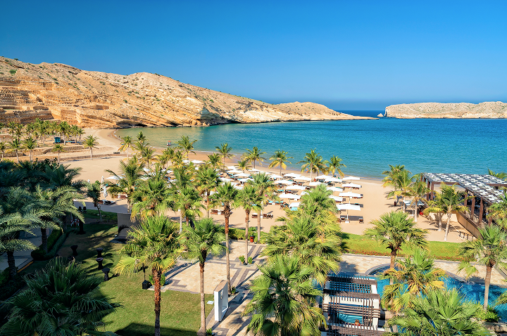 Ein Strand mit einem Pool und einigen Palmen, die sanft im Wind wiegen, unter einem strahlend blauen Himmel.