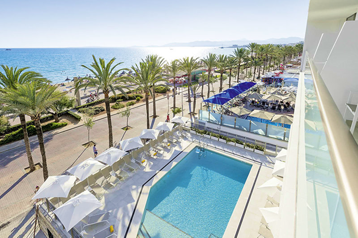 Blick von Balkon auf rechteckigen Pool mit weißen Sonnenschirmen, Palmen und Strand mit Meer im Hintergrund.