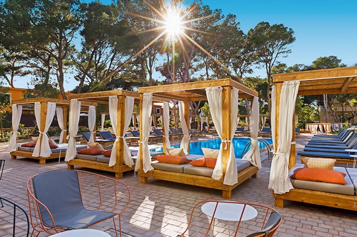 Holz-Pavillons mit weißen Vorhängen und gepolsterten Liegen neben einem Pool unter Bäumen bei Sonnenschein.