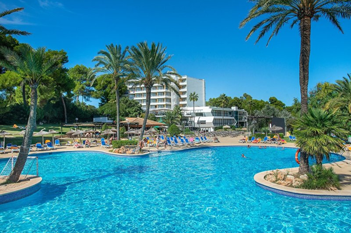 Großer Pool mit Liegestühlen und Palmen vor einem mehrstöckigen Hotelgebäude unter blauem Himmel.