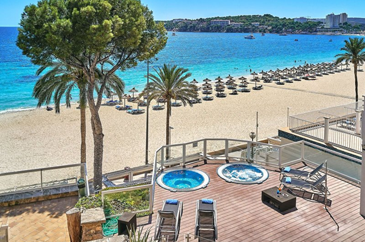 Blick von einer Terrasse mit zwei runden Whirlpools und Liegen auf einen Sandstrand mit Palmen und Sonnenschirmen am Meer.