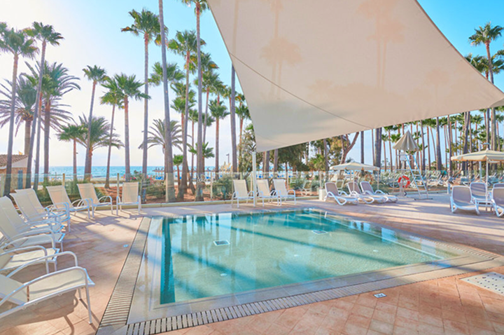 Blick auf den Pool des Strandhotels, umgeben von Liegen und Palmen, mit dem Meer im Hintergrund.