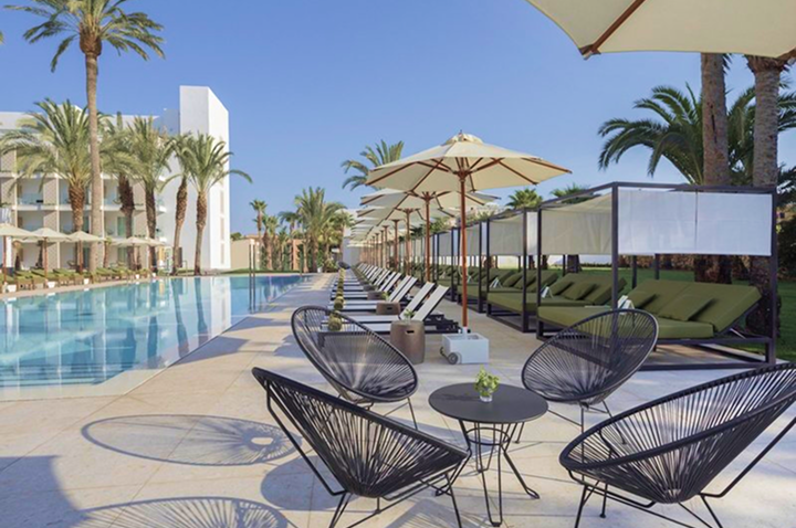 Poolbereich mit Liegen, Sonnenschirmen und runden Stühlen vor einem Hotelgebäude und Palmen.