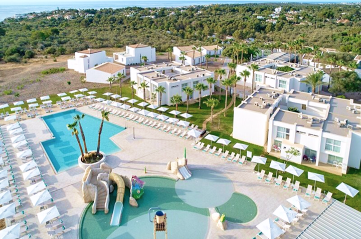 Blick auf ein Hotelresort mit mehreren weißen Gebäuden, zwei Schwimmbecken, Wasserrutschen und Liegestühlen, umgeben von grüner Landschaft.