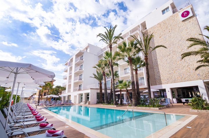 Moderner Hotelkomplex mit mehrstöckigem Gebäude, Palmen und langem Außenpool, links Liegestühle mit weißen Sonnenschirmen und pinken Handtüchern.