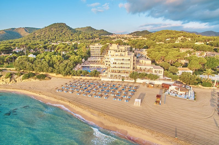 Luftaufnahme eines Hotels mit Sonnenschirmen am Strand und bewaldeten Hügeln im Hintergrund.