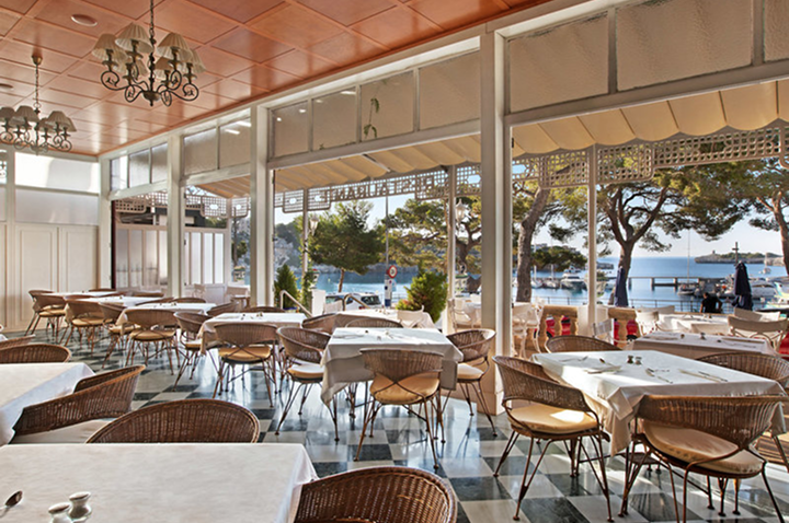 Restaurant mit gedeckten Tischen und Korbstühlen, große Fenster mit Blick auf Wasser und Bäume im Hintergrund.