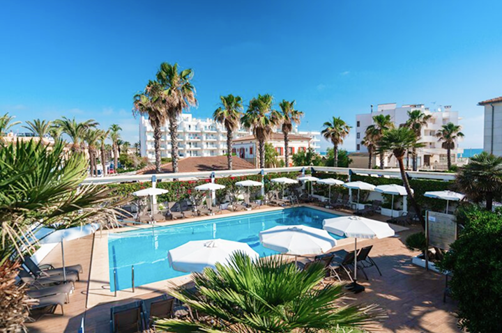 Hotelpool mit mehreren Sonnenschirmen und Liegen, umgeben von Palmen und Gebäuden im Hintergrund.