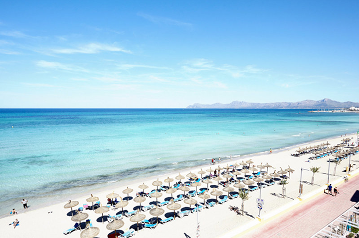 Strand mit Reihen von Sonnenschirmen und Liegestühlen am Ufer eines ruhigen Meeres unter blauem Himmel.