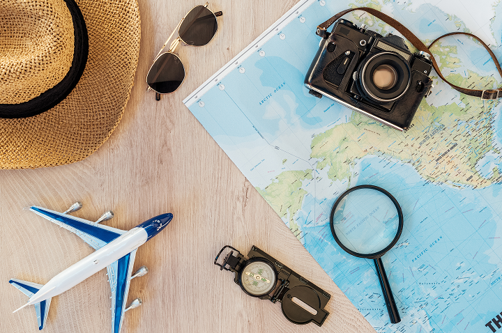 Auf einem Tisch liegen Gegenstände für die Reiseplanung: eine Landkarte, ein Sonnenhut, eine Sonnenbrille, eine Kamera, eine Lupe, ein Kompass und ein Spielzeugflugzeug.