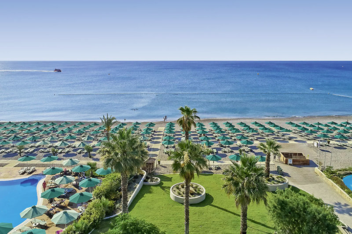 Blick auf einen Strand mit vielen Sonnenschirmen, Palmen und einem Pool im Vordergrund sowie das Meer im Hintergrund.