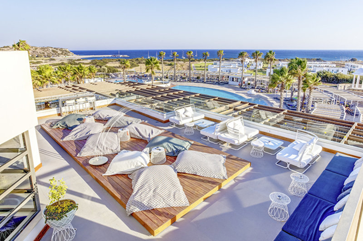 Dachterrasse mit Holzplattform, mehreren Kissen, weißen Stühlen und Tischen, Palmen und Meer im Hintergrund.