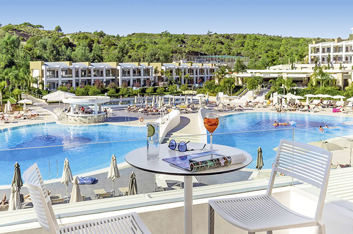 Blick von einer Terrasse mit Tisch und zwei Stühlen auf eine große Poollandschaft vor einem modernen Hotelkomplex mit mehreren Gebäuden und viel Grün im Hintergrund. Blick von einer Terrasse mit Tisch und zwei Stühlen auf eine große Poollandschaft vor einem modernen Hotelkomplex mit mehreren Gebäuden und viel Grün im Hintergrund.