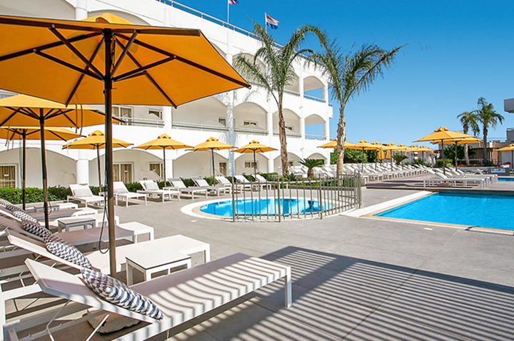 Hotelpool mit mehreren Sonnenschirmen und Liegen, umgeben von Palmen und Gebäuden im Hintergrund.
