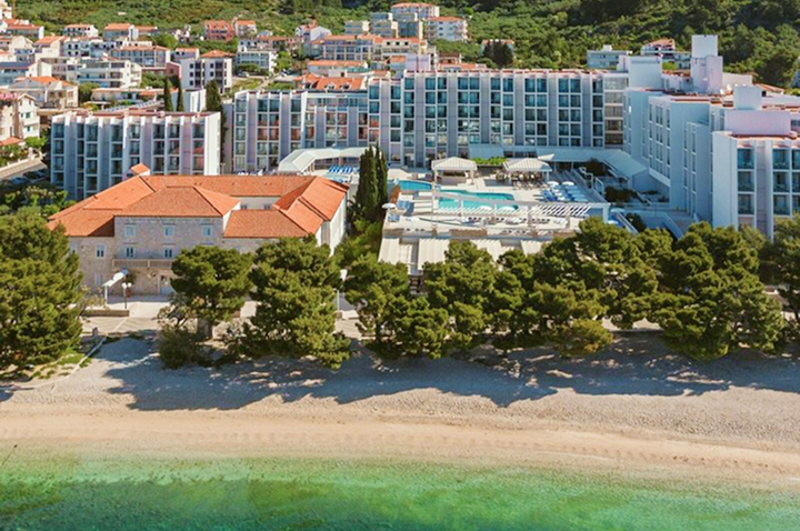 Hotelanlage mit mehrstöckigem Gebäude, Poolbereich und Sandstrand vor bewaldetem Hügel.