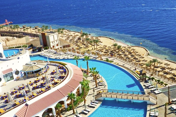 Blick auf den Pool und den Strand des Resorts, umgeben von Palmen und Sonnenliegen.