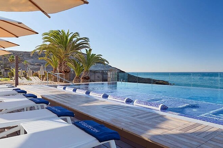 Infinity-Pool mit Holzdeck, Liegestühlen mit blauen Handtüchern und Palmen am Meer unter klarem Himmel.