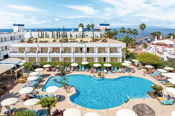 Hotelgebäude mit mehreren Balkonen und einem großen, unregelmäßig geformten Pool umgeben von Sonnenschirmen und Liegestühlen.