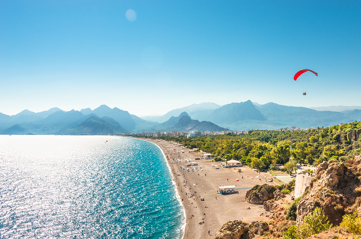 Ein Paraglider schwebt über den Strand und die Berge, umgeben von klarem Himmel und sanften Wellen.