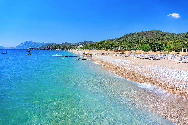 Klarer Strand mit Liegestühlen und Sonnenschirmen neben türkisfarbenem Meer und bewaldeten Hügeln unter blauem Himmel