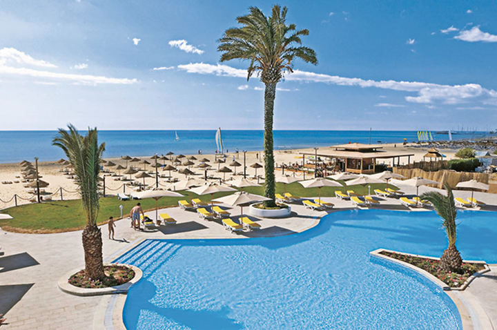 Großer Pool mit Palmen und Liegen, dahinter Sandstrand mit Sonnenschirmen und Meer unter blauem Himmel. Großer Pool mit Palmen und Liegen, dahinter Sandstrand mit Sonnenschirmen und Meer unter blauem Himmel.