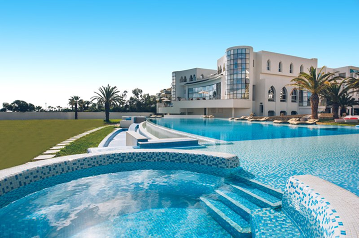 Ein großer Swimmingpool vor einem prächtigen Luxushotel, umgeben von eleganten Gartenanlagen.