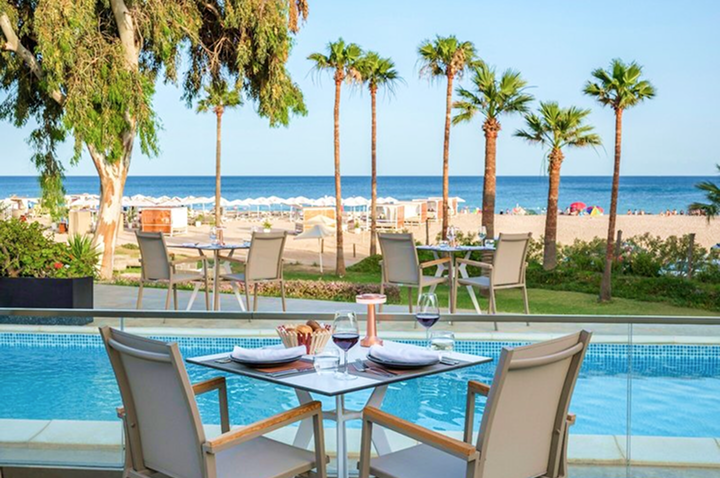 Vier gedeckte Stühle um einen quadratischen Glastisch am Pool mit Blick auf Strand und Meer, Palmen im Hintergrund.