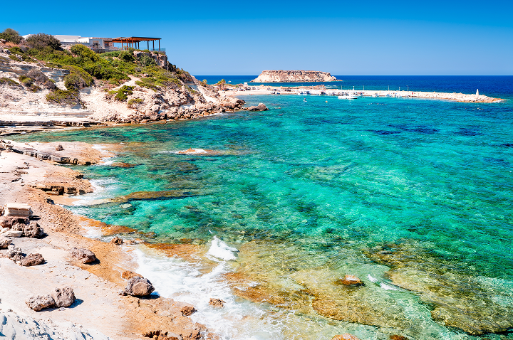 Klares türkisfarbenes Meer an felsiger Küste mit vereinzelten Bäumen und einem Gebäude auf einer Anhöhe unter blauem Himmel. Klares türkisfarbenes Meer an felsiger Küste mit vereinzelten Bäumen und einem Gebäude auf einer Anhöhe unter blauem Himmel.